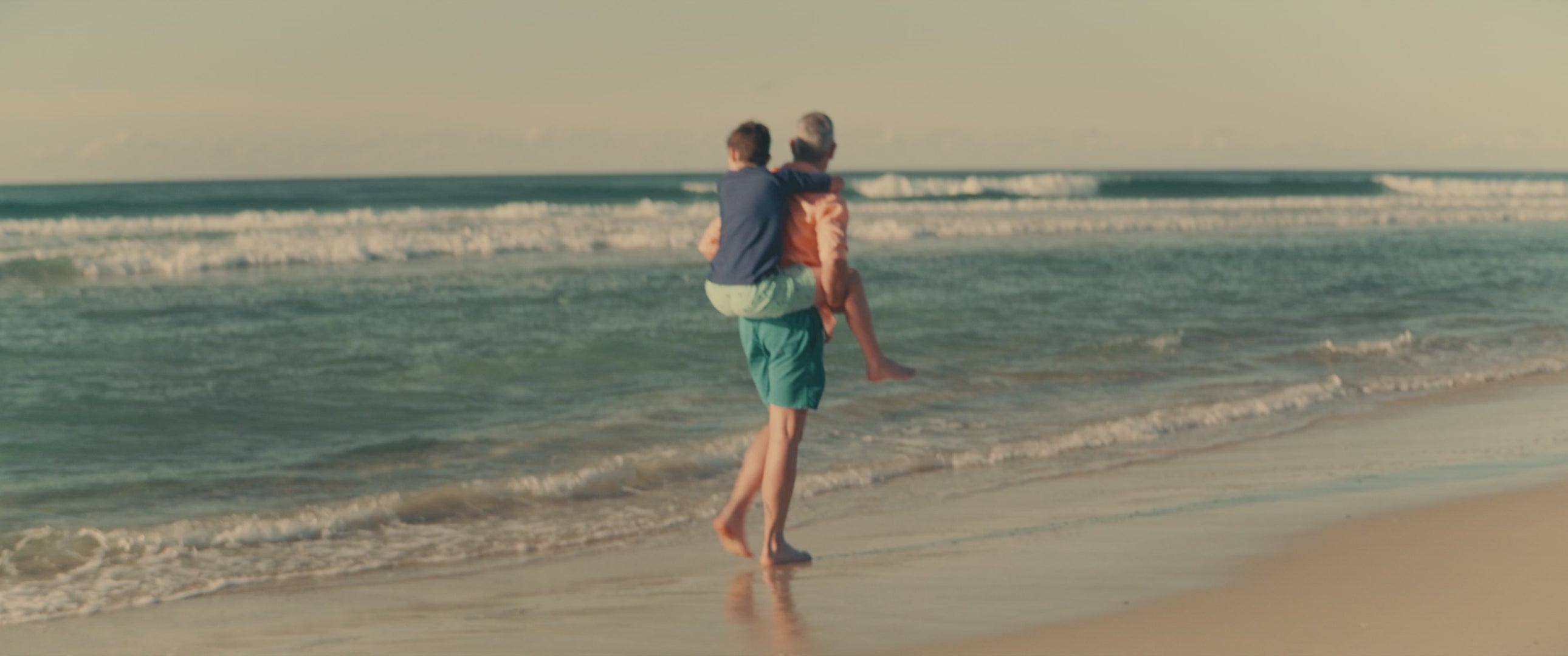 Load video: Father giving his son a piggy-back ride down the beach as the waves gently roll in on a beautiful day.